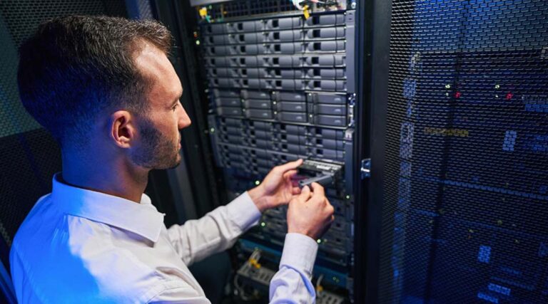 Un homme en chemise blanche installe ou retire un disque dur d'un rack de serveur dans un centre de données, démontrant la définition Infogérance informatique au milieu d'équipements de réseau et d'un éclairage bleu.