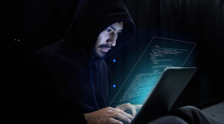 Une personne vêtue d'un sweat à capuche sombre est assise devant un ordinateur portable. Un code holographique lumineux s'affiche dans l'air, laissant présager une activité de ransomware ou des atteintes à la cybersécurité dans une pièce faiblement éclairée.